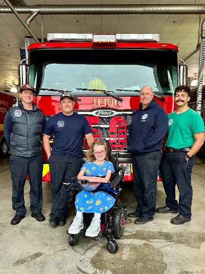 Firefighters standing, a woman in a wheelchair, in front of a firetruck.