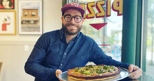 Man in a hat, glases and a beard holding a pizza.
