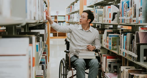 A man in a wheelchair with glasses in a library looking at books.