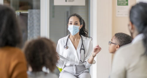 Woman sitting with a mask on and stethoscope around her neck speaking to a group of people.