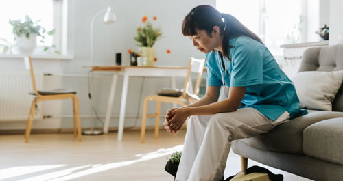 A woman sitting on the edge of a couch looking exhausted and dressed in scrubs
