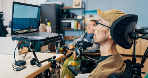 Man in a wheelchair with hat and glasses working on a computer