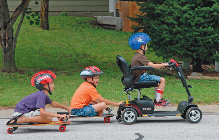 A picture of a little boy in a wheelchair, rolling down a hill with two other little boys on skateboards