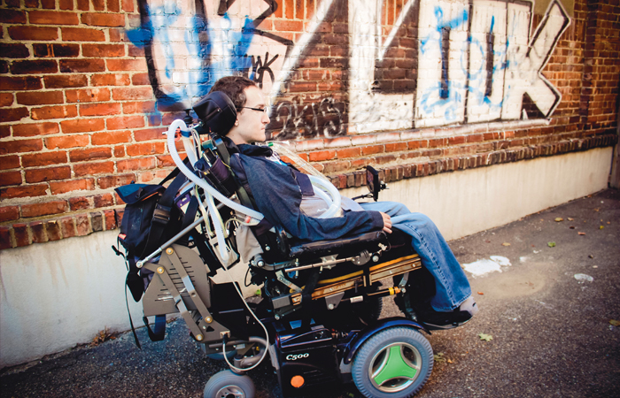 A picture of Jared Aronson. He sits in a wheelchair in front of a brick wall with grafitti on it