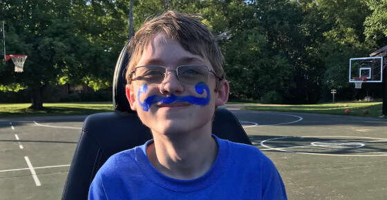 A photo of a little boy in a wheelchair with a makeup blue mustache.