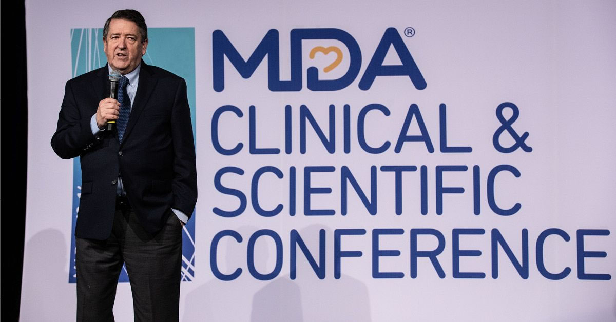 A man, Donald S. Wood, PhD, President and CEO of MDA standing with a microphone with his hand in his pocket in front of the MDA Clinical & Scientific Conference banner