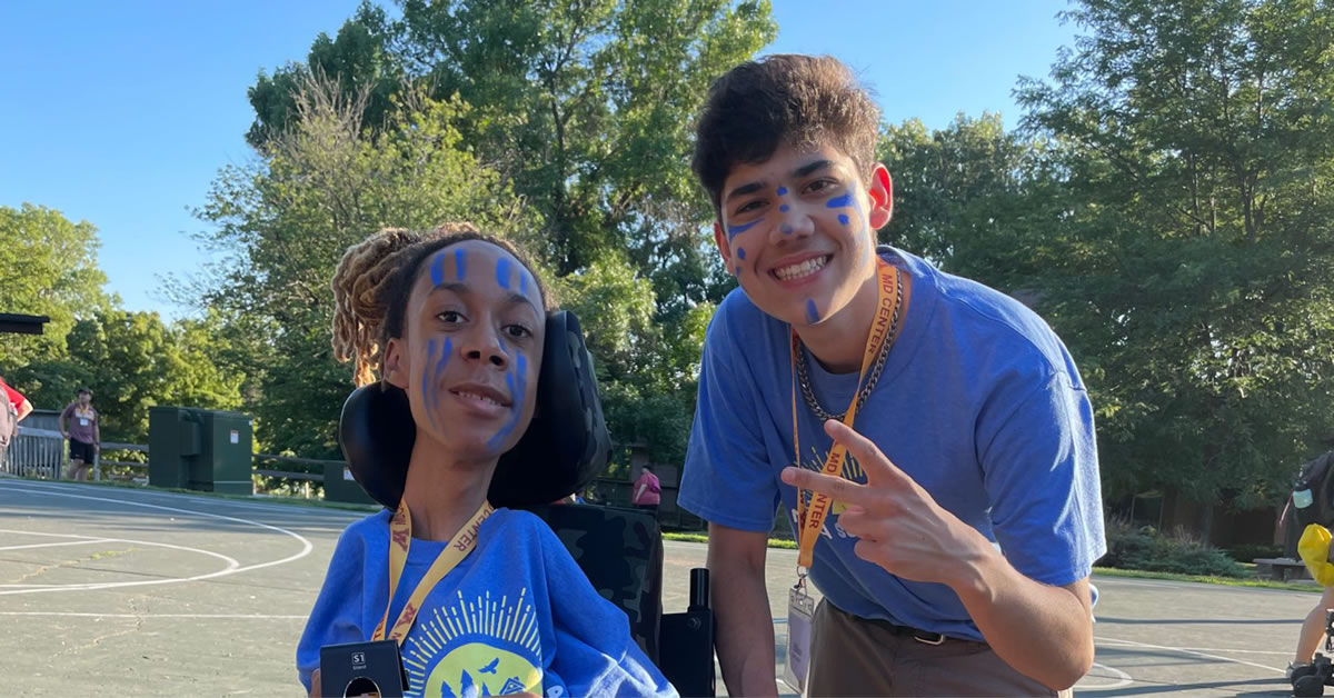 A girl in a wheelchair smiling and a boy bending over smiling while giving the peace sign. 