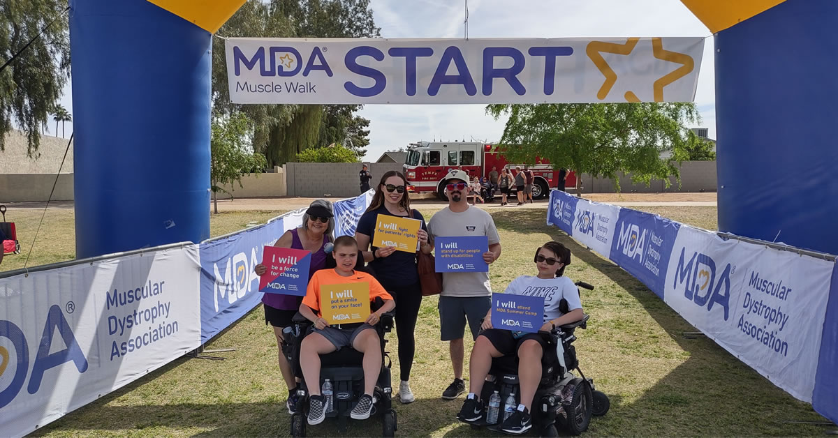 People standing and posing for a picture in a grassy area under an MDA Muscle Walk Start banner and arch. There is a firetruck in the background.