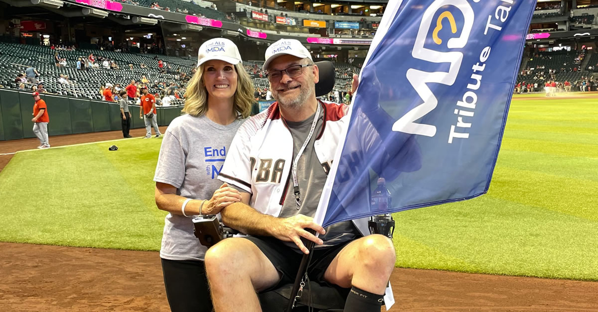 Woman standing next to a man in a wheelchair inside of a baseball stadium. The man holds an MDA Tribute flag