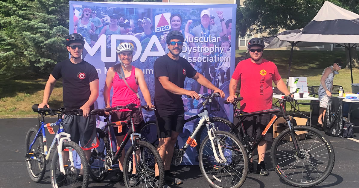 Four people standing next to their mountain bikes in front of a MDA banner. Table tent to the right and trees in the background.