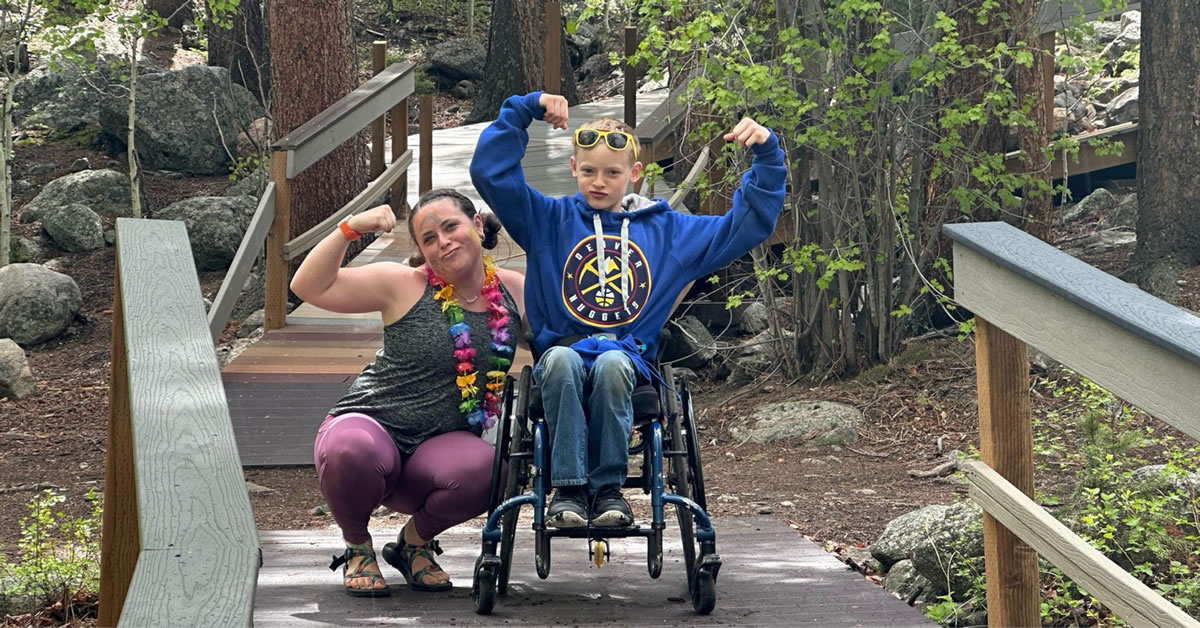 Image of a woman and boy flexing their biceps on a wooden path in the woods. The woman is squating down and the boy is sitting in a wheelchair.