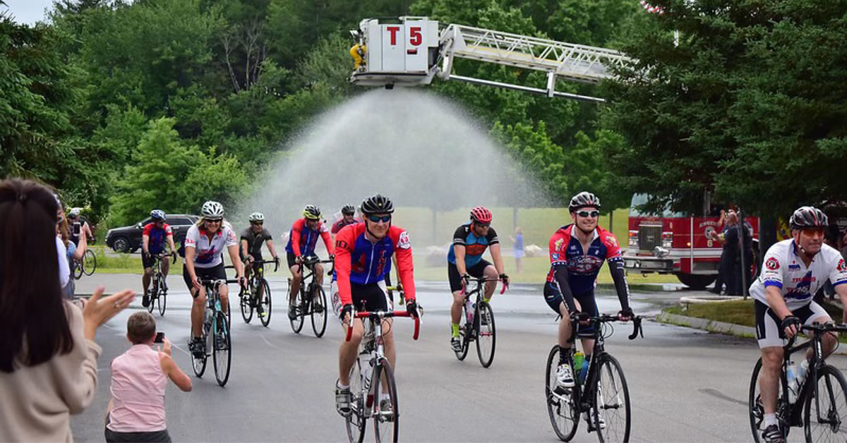 People on bicycles riding in a street past a fire truck and fans.